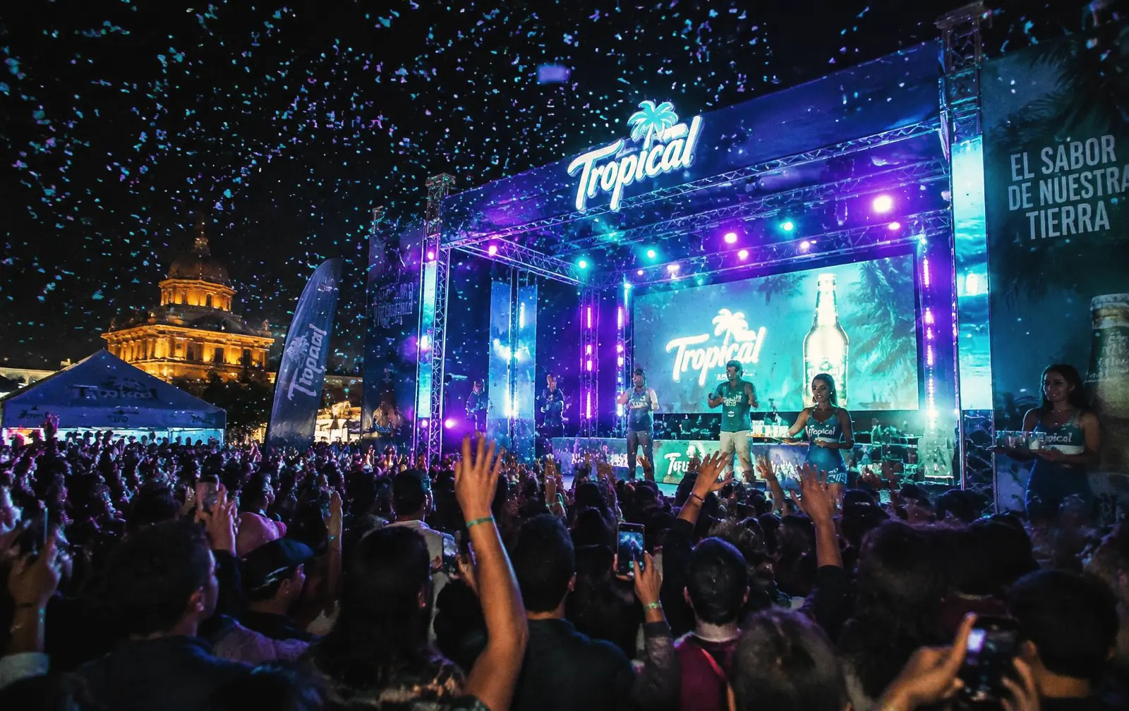 Escenario de una activación nocturna con luces moradas y turquesa, confeti en el aire y público con las manos arriba