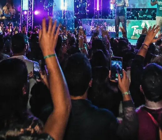 Manos del público levantadas frente a un escenario, una sostiene un teléfono grabando